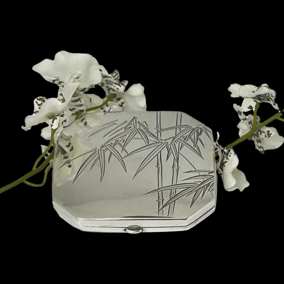 Main photo of Oriental 950 silver compact seen from a slight height looking diagonally down at the decoration on the lid cover. There are 2 bamboo stalks slightly on the right side of lid with the bamboo leaves over the top and one spray of leaves hanging over to the left hand side. The compact is set against a black background with a spray of white orchids placed around it for display purposes.