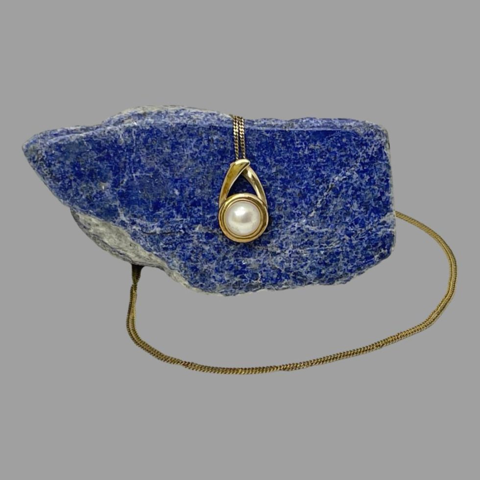 Main photo of gold pearl pendant &chain with the pendant hanging off a side of natural lapis lazuli rock and the chain laid across the top of the rock and extended out from the left and laid in a circular form around the stone. The gold setting for the pearl is on a circular design which has 2 short arms, one above one below like a piece of string laid with one circle loop and the end of the string laid above it. The ends of the gold 'string' acts as the bail for the chain - very neat!