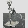 Main photo of Dutch street lamp lighter displayed on top of a block of black marble. The miniature is posed at a diagonal angle to show most areas of the piece. The gas street lamp is to the left foreground, the ladder is leaning against it on other side with the workman either ascending or descending the steps. His right hand is holding onto the ladder whilst his left hand appears to be holding something.