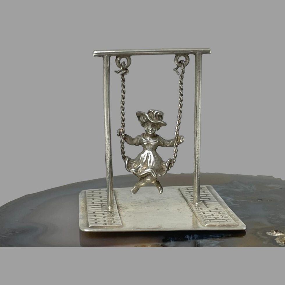 Main photo of miniature girl on swing seen from an eye level with the girl forward facing. The piece is displayed on top of a slice of natural agate. This is an articulated piece with the swing actually swingable! Lovely desk toy gift.