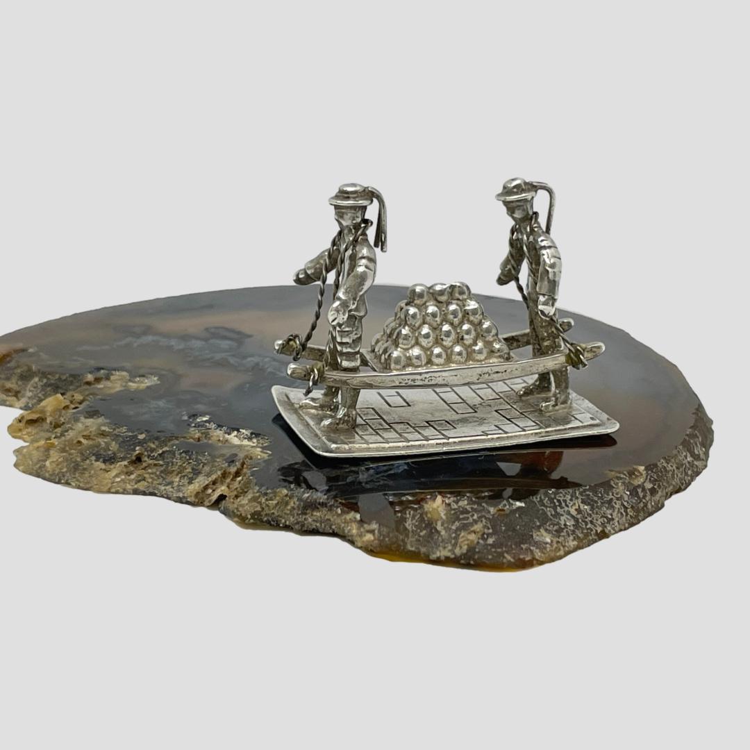 Main photo of Dutch miniature cannonball carriers seen from a side angle with the carriers facing left and the cannonballs neatly stacked up on a barrow. The men look like they should be holding the ends of the barrow but they are just carrying it around their necks. Perhaps punishment of sorts ? Lovely details to the sailors uniforms and hats and again, the cobbled streets of Holland. The miniature is displayed on top of a slice of natural agate.