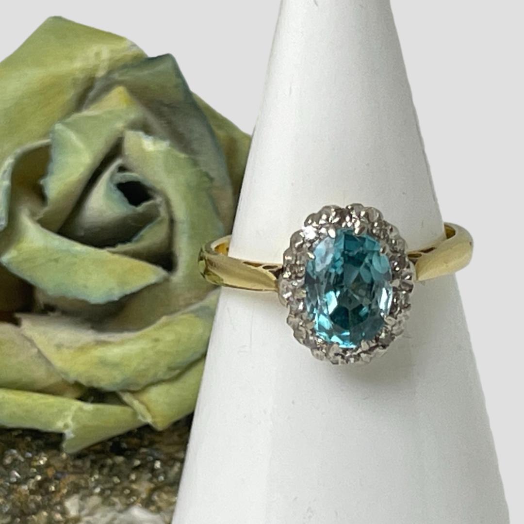 Main photo of gold blue zircon ring displayed on a cone shaped stand and displayed with the ring front forward facing. The blue zircon is an incredible colour like turquoise crystal and the cut of the stone picks up the beauty of the gem perfectly. The 10 small round cut diamonds framing the zircon are set in 18 karat white gold while the band is 18 karat yellow gold. A blue rose which is more green is seen to the back left of the ring stand.