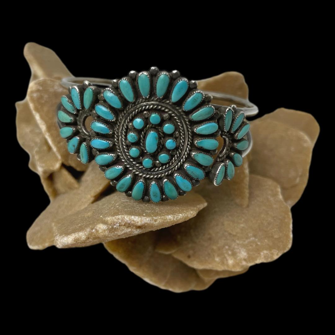 Main photo of Southwest needlepoint turquoise cuff displayed on top of a rock of natural desert sand rose with the bracelet front on show. The cuff has 3 sections of turquoise; the central area is the largest. This area is circular with a single pine nut shaped turquoise set vertically at the centre. Around this, there are 8 small round turquoise stones followed by a section of silver rows - twist, plain, twist then 21 small petal shaped turquoise pieces laid in a circle with pointed ends on the inside. To either side of the central circle are smaller half circles with open end on the inside. These areas are decorated with 7 petal shaped turquoise pieces with the pointed ends on the inside. The colour of the turquoise is a slightly darker sleeping beauty shade with a few of the stones showing a little more green.
