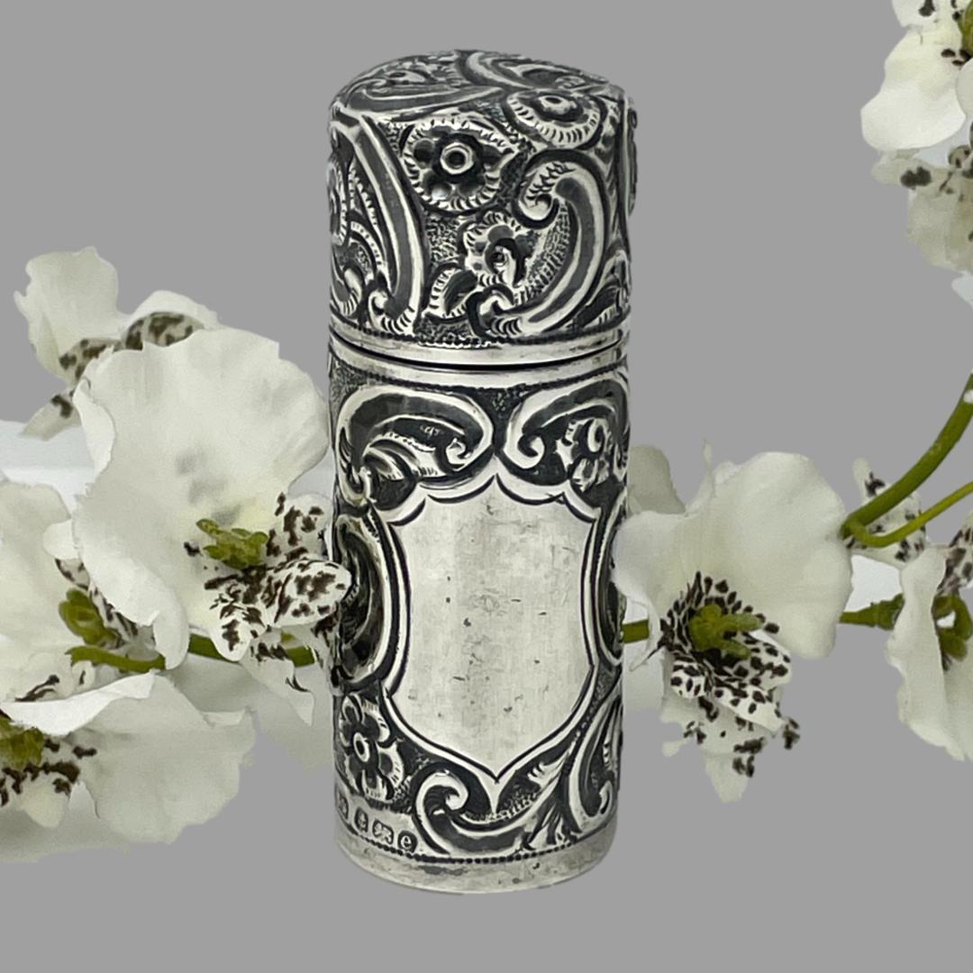 Main photo of portable silver scent bottle displayed on a flat surface with the empty cartouche side forward facing. The bottle has some very intricate embossed work throughout mostly swirls with the occasional flower. The full hallmark is just visible to the bottom left. A spray of white orchids lays behind the bottle for display purposes