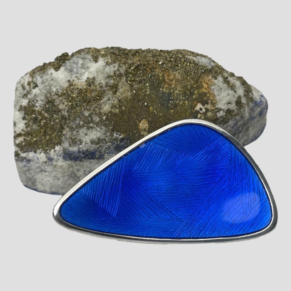 Main photo of Norwegian blue enamel brooch displayed leaning against a natural rock with gold flecks with the enamelled front on display. The shape of the brooch is like a set square ruler but not 90 degree angle! It also has a light concave design.