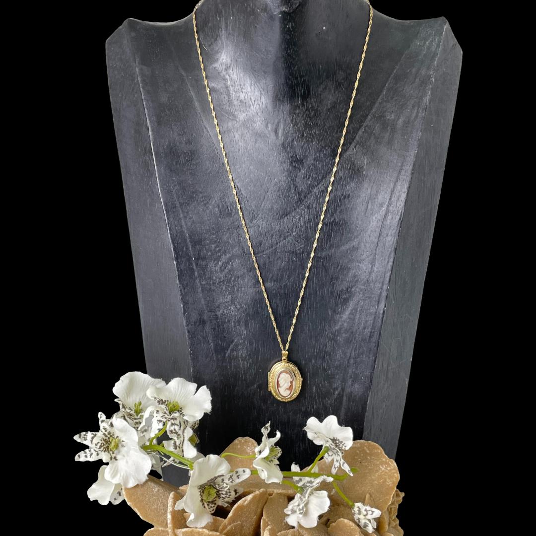 Main photo of gold cameo locket & chain displayed on a black wooden display stand and seen with necklace front forward facing. The chain is a generous length at 460mm long and compliments the locket perfectly. There is a rock of desert sand rose with white orchids on at the base of the display which has colours similar to the cameo.