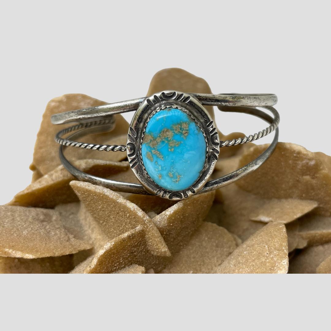 Main photo of Native American turquoise cuff displayed on top of a rock of natural desert sand rose and shown with the front forward facing. The turquoise is a beautiful shade and has some natural golden coloured inclusions. The stone is framed in sterling silver with the cuff band in 3 rows; 2 plain at top and bottom and a twisted one at the centre. These gather at the end of the cuff.