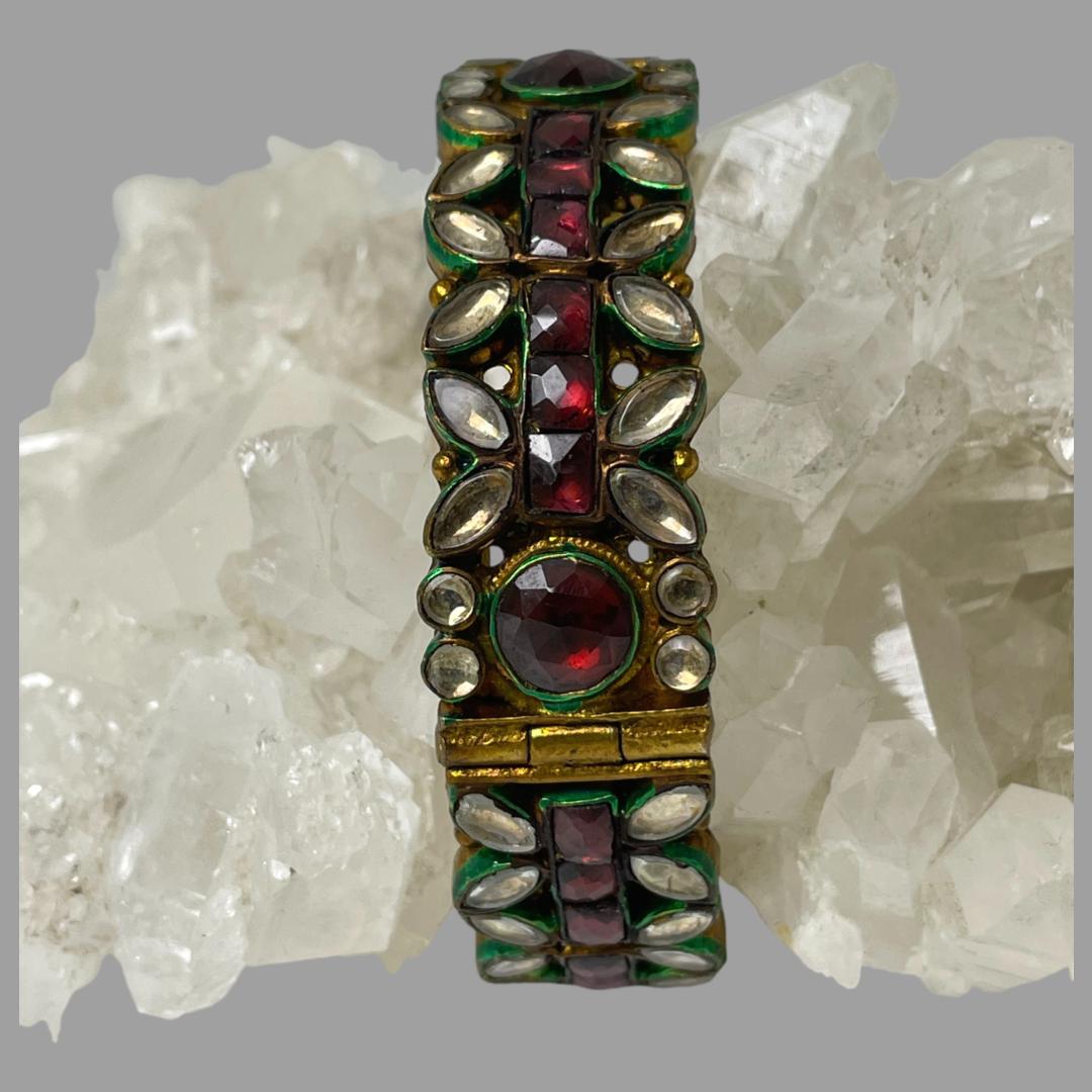 Main photo of old Indian glass bracelet displayed around a rock of natural quartz. The bracelet is constructed from a variety of glass beads; square cut ruby red beads make the central line with a large round red bead at the hinged area. There are almond shaped clear glass beads on the outer edges either side of the central red pointing inward and 2 small round clear glass beads either side of the large round red bead. There is green enamel around each bead and the main base is unmarked silver gilt.