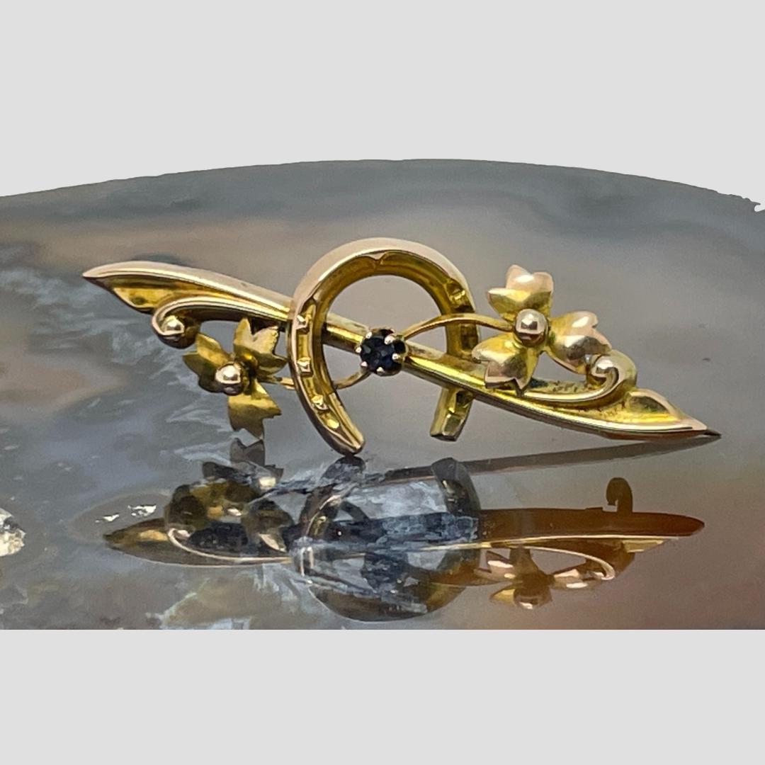 Main photo of Edwardian gold sapphire brooch displayed forward facing on top of some natural agate. The horseshoe is in the centre with the pin bar going through it. A single round cut sapphire sits in the middle. A twig with leaves or maybe flowers on either end sits across the pin bar and behind the sapphire so the pin bar and twin crisscross in the middle. The pin is open and propping the brooch upright.