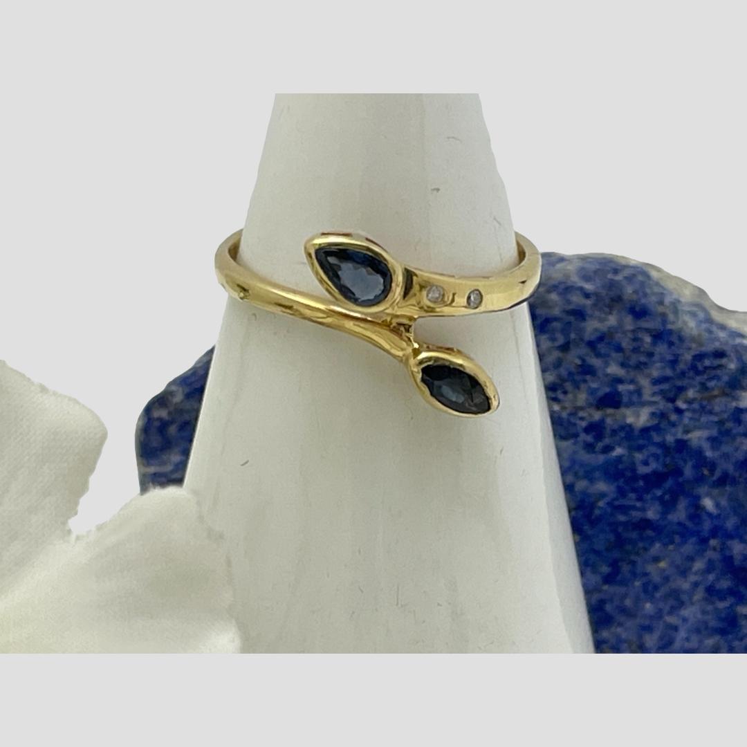 Main photo of 18 karat gold sapphire and diamond ring displayed on a cone shaped stand with the front forward facing. A small rock of lapis lazuli is behind the cone stand and petals of a white flower on the left.. A lovely split band ring with pear and drop cut sapphires to either end. The 2 small round cut diamonds are visible in the shoulder of the top band. Although it has a split band, the ring is not adjustable.