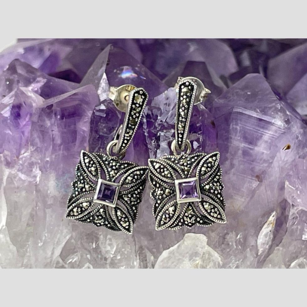 Main photo of deco style silver marcasite earrings displayed on a rock of natural amethyst crystal and shown forward facing. The top part of the earrings is line of silver with marcasite with square attached at ends housing a small square cut amethyst to centre of each within a frame of marcasite stones.
