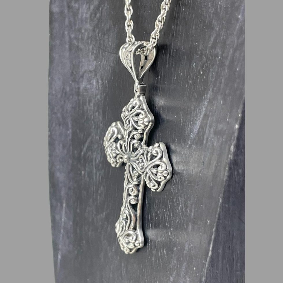 Photo showing necklace hanging from a display stand with only the cross area on display. The photo is taken from a slight side angle with the cross facing left. Intricate pierced silver work side on display.