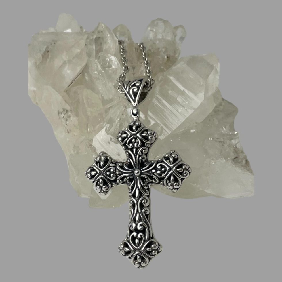 Main photo showing the cross leaning against a rock of natural quartz crystal. The side displayed is the more intricate pierce worked side and most likely the front The bail is as intricately pierced as the cross and part of the chain is visible to the top laid on the crystal.