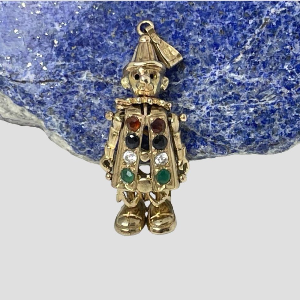 Main photo of the articulated clown pendant leaning up against a piece of natural blue lapis stone and facing forward. The clowns coat has gemstone buttons starting with red then blue clear and finally green.