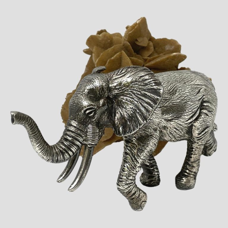 Main photo of elephant posed in front of a rock of natural desert rose. The elephant is posed slightly at a diagonal angle with the raised trunk in the bottom left. This elephant is a bull with fine tusks.