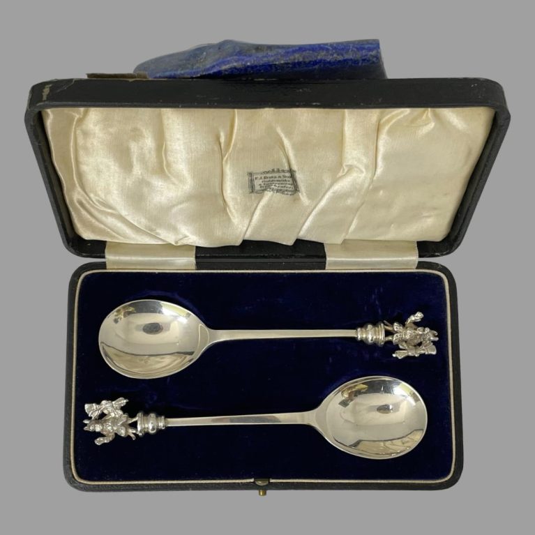 Main photo of silver trusty servant spoons spoons displayed inside their fitted case and seen looking straight down from above. The lid is leaning upright against some lapis rock.