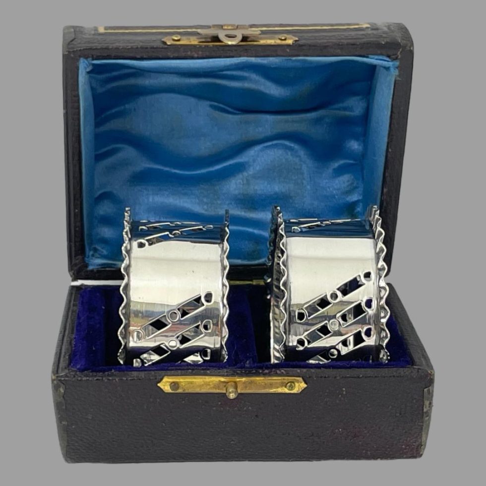 Photo of the sterling silver napkin rings inside their fitted case and seen with box front forward facing and the empty cartouches on show. The blue satin inside lid is shown.