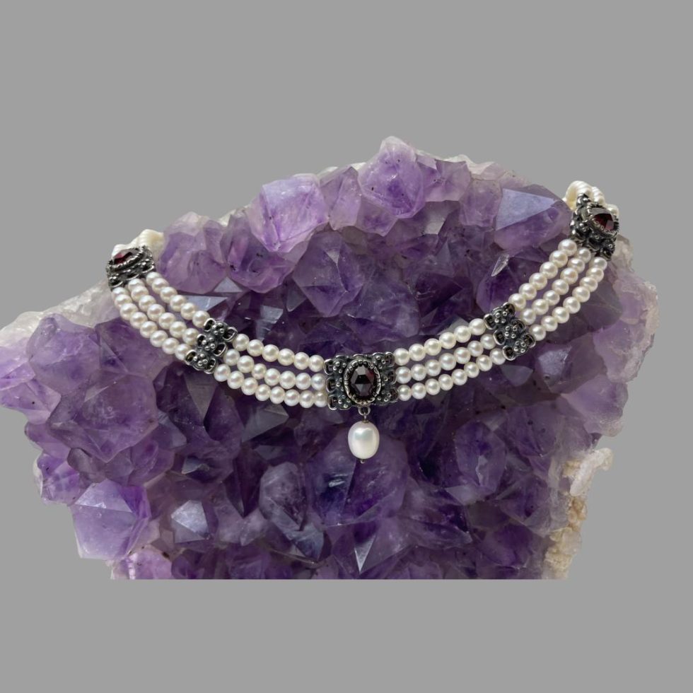 Main photo of 835 silver pearl and garnet necklace draped on a piece of natural amethyst rock. The rock is upright and the necklace hangs in a wide U shape. The centre part of the necklace has a single drop pearl hanging below the garnet. Next area of silver on either side of the central piece is thinner and is just plain silver and the final 2 sections either side of the thin area also houses a garnet but without a hanging pearl. 3 rows of pearls go all the way round. This is Princess making jewellery!