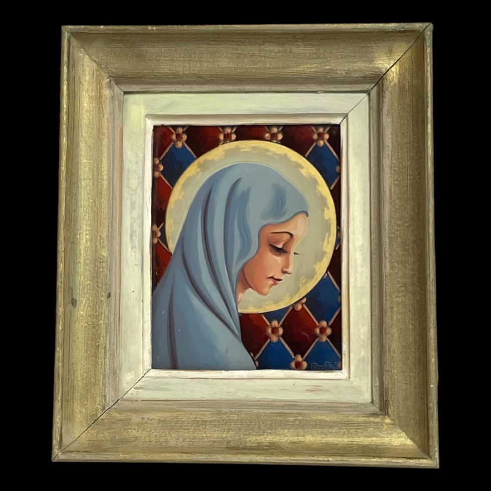 Main photo of the French reverse painted glass panel showing the whole panel and frame. The frame is a light golden colour. The Virgin Mary is wearing a light blue habit, face in profile facing downward and to the right and she has a feint smile. Her halo could be a full moon behind her head and the background is a patchwork of red and blue diamonds separated with small flowers. Artists signature to bottom right of panel.