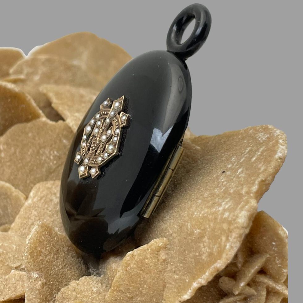 Photo of Victorian jet locket displayed on top of a rock of natural desert rose with the monogrammed locket front facing left. The hinge side of the locket is also visible.