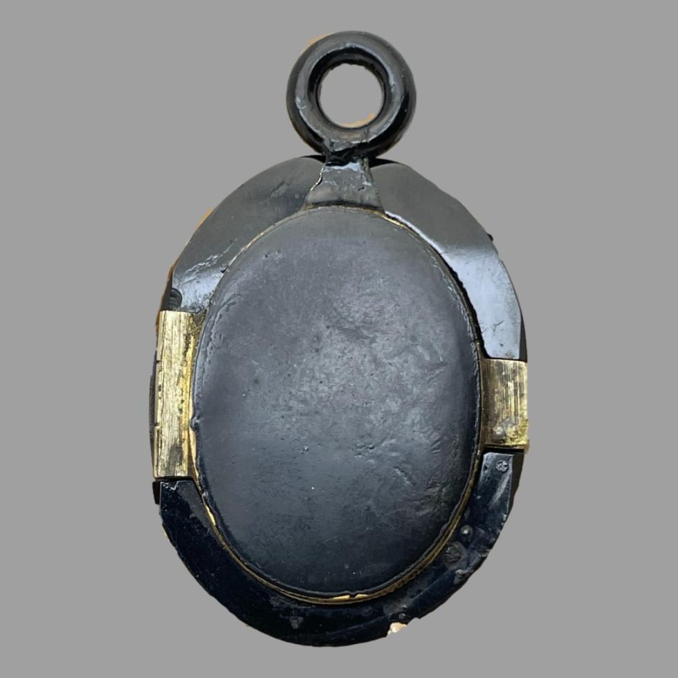 Photo of the back of closed Victorian jet locket showing the unpolished jet and the brass hinge to the left and opener to the right. The back section is smaller than the front of locket.