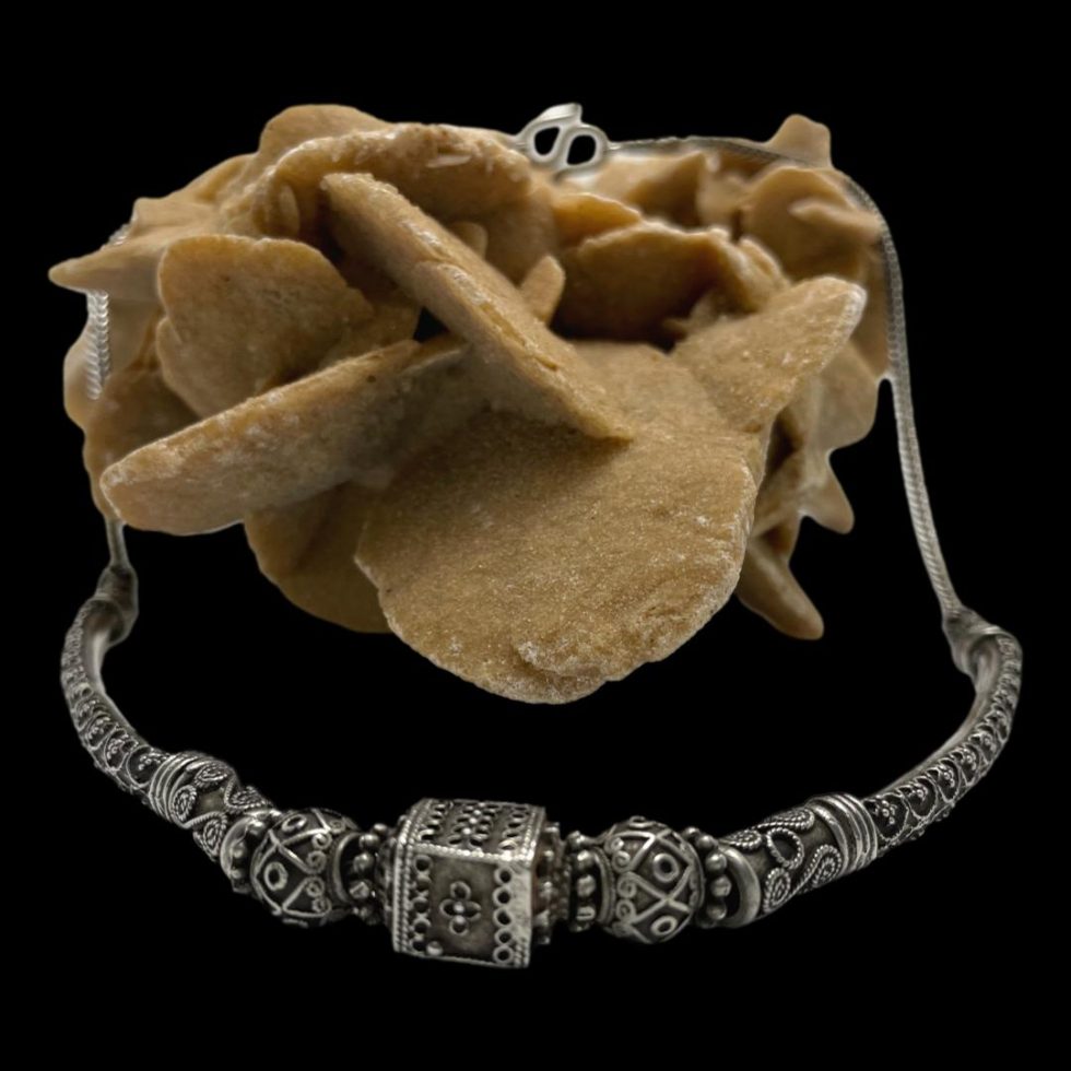 Main photo of vintage Rajasthani silver necklace displayed draped over a rock of natural desert sand rose. The front end of the necklace with the intricately embossed beads is in the foreground whilst the clasp area is on top of the desert rose rock. The centre bead is square with round beads either side followed by small tubular beads then finally a longer tapering bead before snake chain. The clasp ends of the necklace also have embossed bead detail.