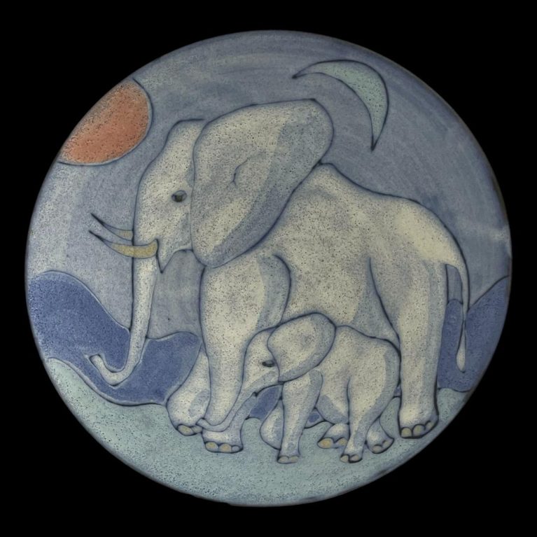 Main photo of Tessa Fuchs elephants bowl seen looking straight down into the bowl centre looking at the whole detail of the elephants adorning this piece of studio art. The sun is red and to the top left and crescent moon is blue and to the top right. The elephants are facing left towards the red sun. The elephants are a pale grey blue with blue areas to highlight the shadows. The adult elephant takes up quite a large portion of the picture. The calf walks beside the adult and fits between its front and back legs. The ground, mountain range in background and sky are different shades of blue - the darkest being the mountain range.