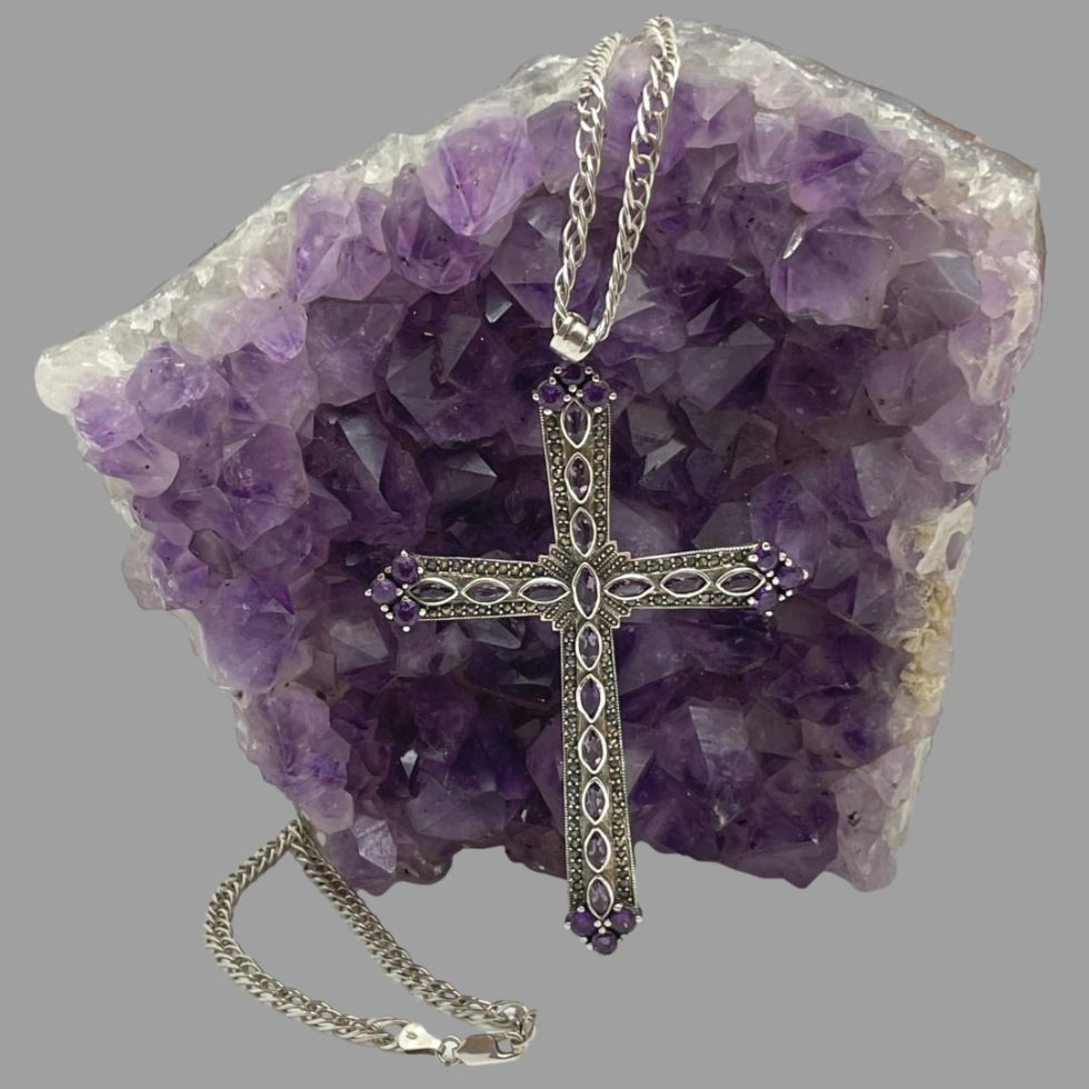 Main image of the large silver amethyst cross and chain with the cross leant up against a rock of amethyst crystal and the chain extended up and over the rock and reappearing from the lower left of rock with the clasp ends in the foreground.