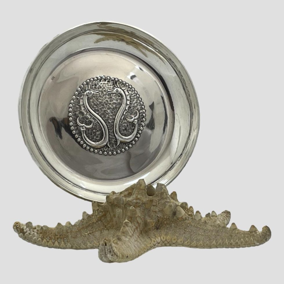 Main photo of sterling silver seahorse dish shown displayed on top of a natural starfish (doing the sea theme here!) The dish has a central area which is hand chased with a stylised seahorse design of 2 seahorses facing each other in mirrored form.. The area outside of the hand chased centre is smooth silver with a shallow lip around the edge.