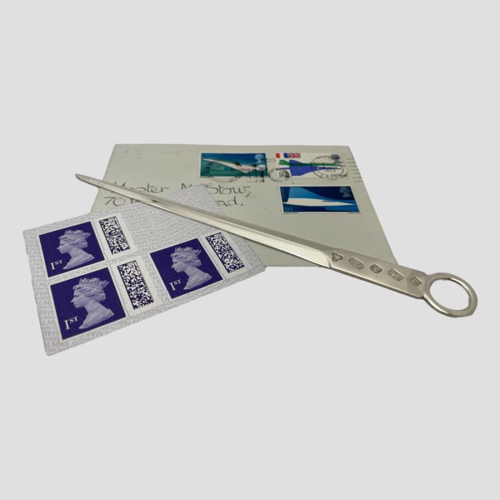 Photo of cased silver letter opener shown with letter opener outside of its case and displayed across an old stamped envelope and some present day stamps.