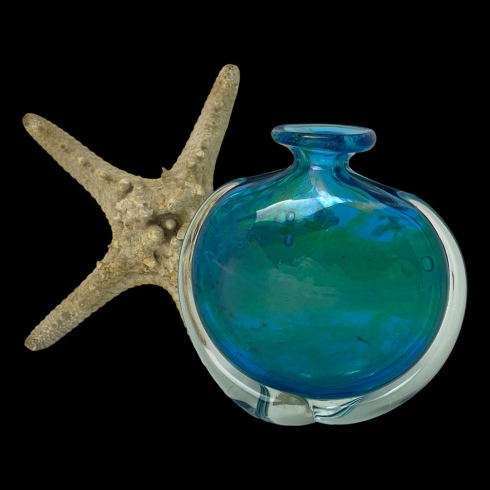 Main photo of unsigned Mdina glass vase see with one flat side facing forward. The clear glass ring around the bulbous area is visible with the beautiful blue and ochre glass sitting on top of that. There is a starfish displayed behind the vase to the left to give added seaside feel.