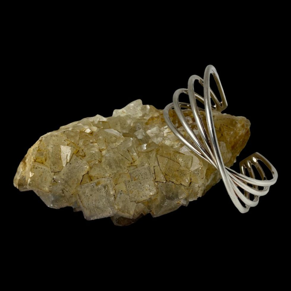 Main photo of Hans Hansen silver cuff displayed propped onto the end of a rock of natural river quartz with the twist front displayed in the foreground. The cuff bracelets design is quite unique appearing like a bow of silver bands.