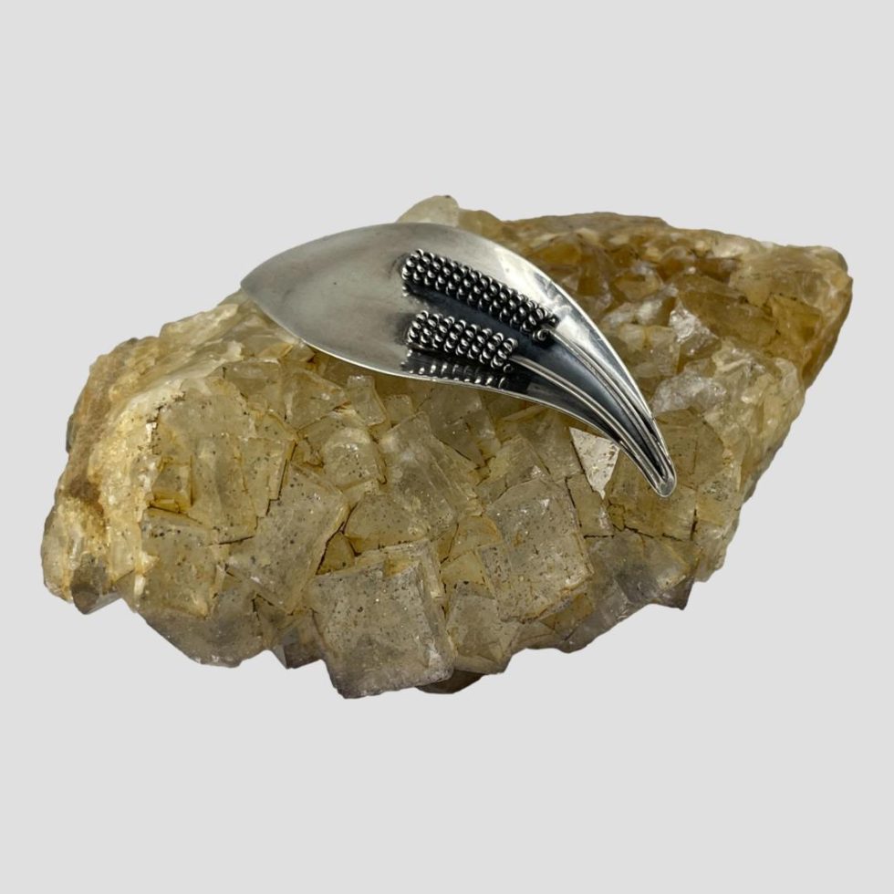 Main photo of Danish silver calla lily brooch displayed on top of a rock of natural river quartz. The pointed end where the stamen begin from is pointed down to the bottom right corner whilst the top of flower is facing towards top left.