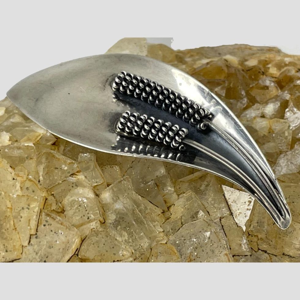 A close up photo of the front of Danish silver calla lily brooch showing the marriage of simplistic and intricate design. The main body of the flower is smooth silver sheet in a tear drop shape while the 2 stamen have fine detail to the anther to produce the look of pollen attached.