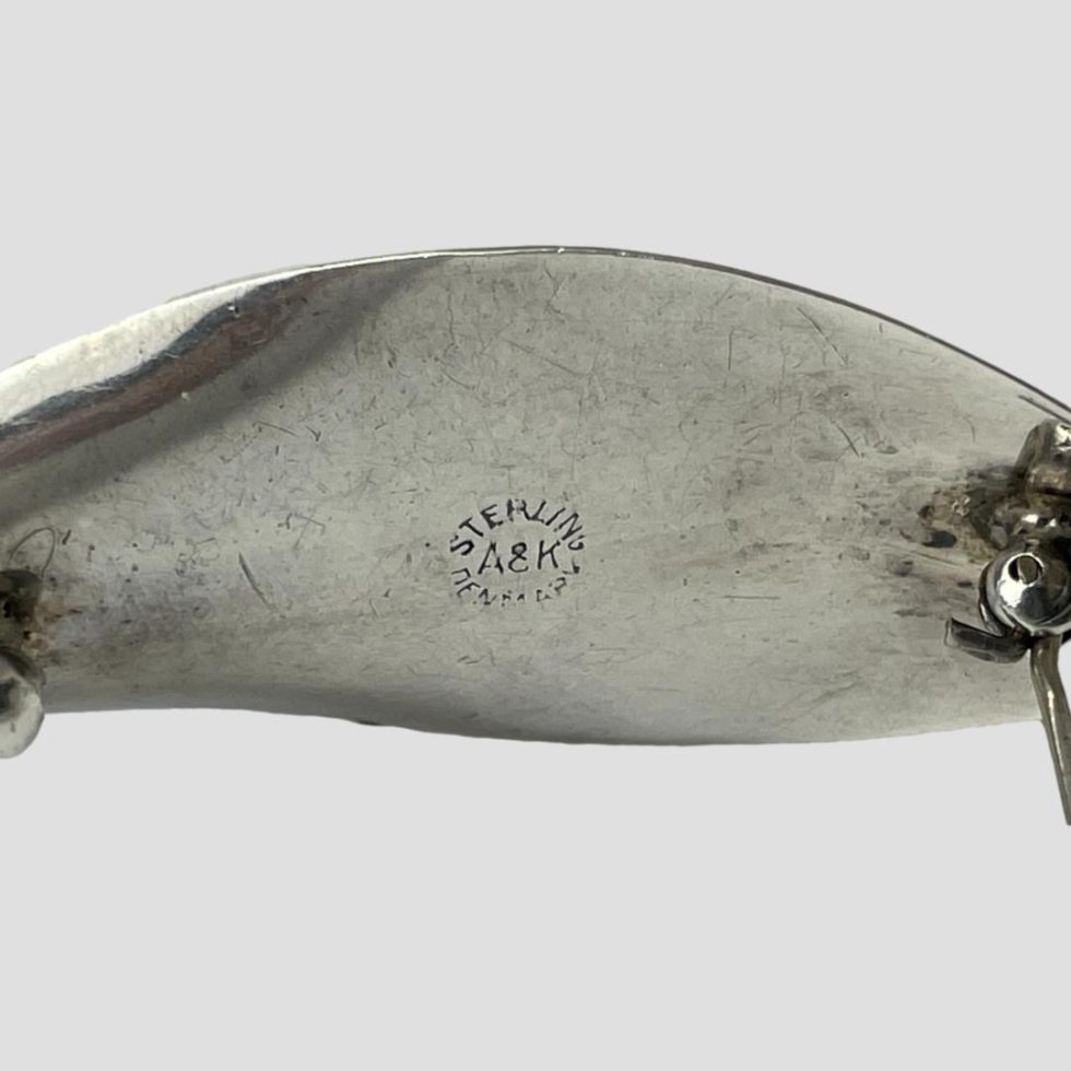 Close up photo of the back of Danish silver lily brooch. The pin is open and the hallmark is on display reading Sterling in semi circle at top Denmark in semi circle below and A&K to the centre. The brooch is still on top of a rock of natural river quartz.