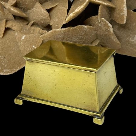 Main photo of Victorian brass vesta box seen with top closed and the box frontage facing very slightly off to the left. The box is basically shaped like a trunk and is of good solid quality. The feet are chevron shaped although this is only visible from the base and protrude a little from the box base. The box is displayed in front of a rock of natural desert sand rose which is partially reflected in the box lid.