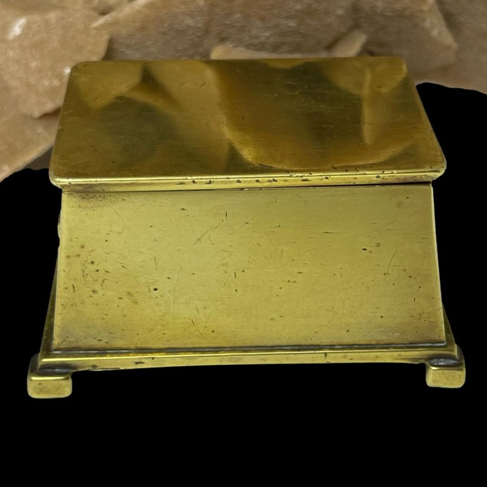 A close up of the Victorian brass vesta box seen from the front opening side with lid closed. The desert sand rose is still in the background and reflecting onto the lid.