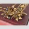 Main photo showing the 9 karat gold multi gemstone floral spray brooch displayed forward facing on the corner of a burgundy coloured old book. The brooch is laid across the bottom corner of the book cover which has a pretty gilt decoration. There are 3 floral blooms; top 2 with garnets to the centre and lower one with sapphire. There are 4 buds, 2 of emerald, 1 ruby and 1 sapphire. The flower petals and spray are in 9 karat gold.