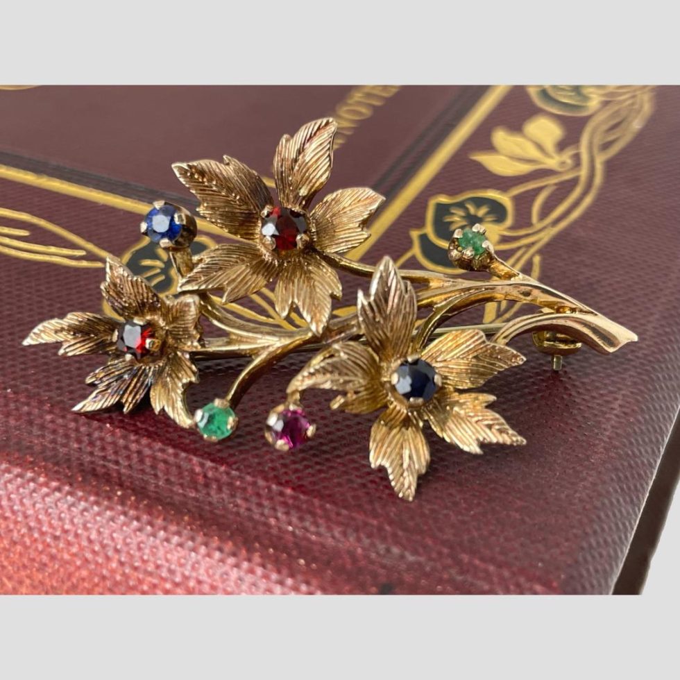 Main photo showing the 9 karat gold multi gemstone floral spray brooch displayed forward facing on the corner of a burgundy coloured old book. The brooch is laid across the bottom corner of the book cover which has a pretty gilt decoration. There are 3 floral blooms; top 2 with garnets to the centre and lower one with sapphire. There are 4 buds, 2 of emerald, 1 ruby and 1 sapphire. The flower petals and spray are in 9 karat gold.