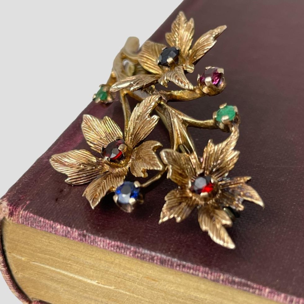 The multi gemstone floral spray brooch is still displayed on the corner of a burgundy coloured old book and seen this time from a diagonal side angle with the smallest of the 3 flowers with garnet to centre in the foreground.