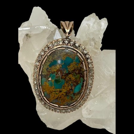 Main photo of the 9 karat gold turquoise and diamond pendant displayed hanging from a rock of quartz crystal. The pendant is forward facing. The natural turquoise stone is a generous size and is not a purely turquoise stone - it has inclusions of natural rock that the turquoise was in. There is a small missing piece of the stone to the middle left which adds to this pendants charm rather than diminishes it.