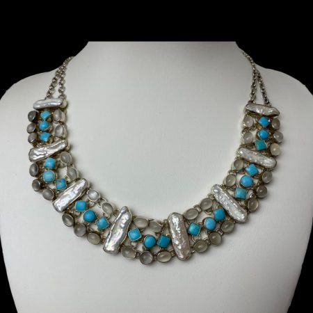 Main photo of the moonstone, turquoise and pearl collar style necklace displayed on an off white display stand and forward facing. The necklace sits in a wide U shape and can be worn either on the collar bone or just below. There are 8 segments, each separated with a vertical baroque pearl. In between the pearls are 3 rows with 2 horizontal oval moonstones at top followed by 3 turquoise stones-the 2 outer stones are square and centre one is round and finally a last row of more moonstones - 2 horizontal oval at ends and round in centre. The whole ensemble is fixed with 2 chains at either end which tapers to one. There is something very magical about this necklace as if it was made for the Empress of the Sea or Moon Goddess.