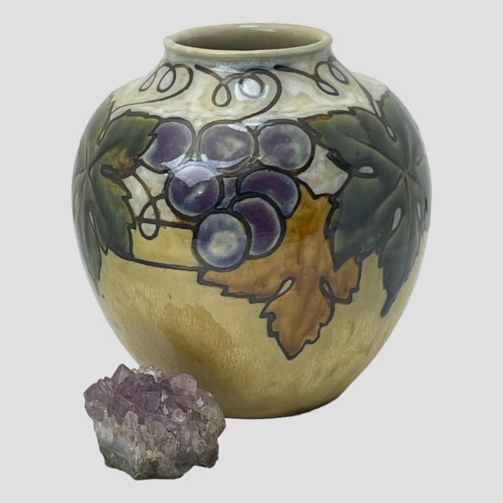 Main photo of Ethel Beard grapevine vase for Royal Douton seen from an eye level. The base colour of the vase is a pale mustard on the lower end and a greyish cream at the top. The decoration on display in this photo is 2 dark green vine leaves partially visible on the left and right with a small bunch of purple grapes near centre of the leaves and a burnt orange colour leaf lower down right of grapes with lines of the vine visible. There is a small rock of natural amethyst crystal to the bottom left of vase which could be a small bunch of grapes with quite some stretch of the imagination!