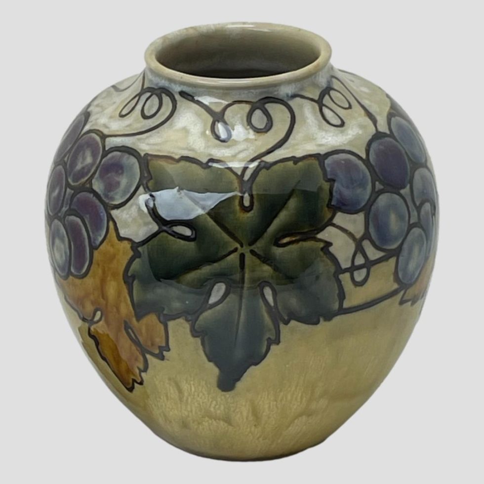 Photo of vase seen this time with a whole dark green vine leaf at the centre and partially visible grapes on the left and right. The burnt orange leaf is visible to the lower left. The angle of the photo is slightly raised peering just into the mouth.