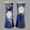 Main photo of Tall Doulton rose vases seen side by side from an eye level. The vase on left is displaying the side with rose lower down and the one on right has rose at the top. The background colour of the vases is a rich royal blue with one large off white rose to each with a few rose buds in a brown colour and leaves in a mixture of green and light brown. The rim of the vases is a creamy white. The vases can be displayed like this (one rose higher, one lower) or with both showing roses at same height. The vases have a lovely chimney-like shape and are stunning even without flowers!