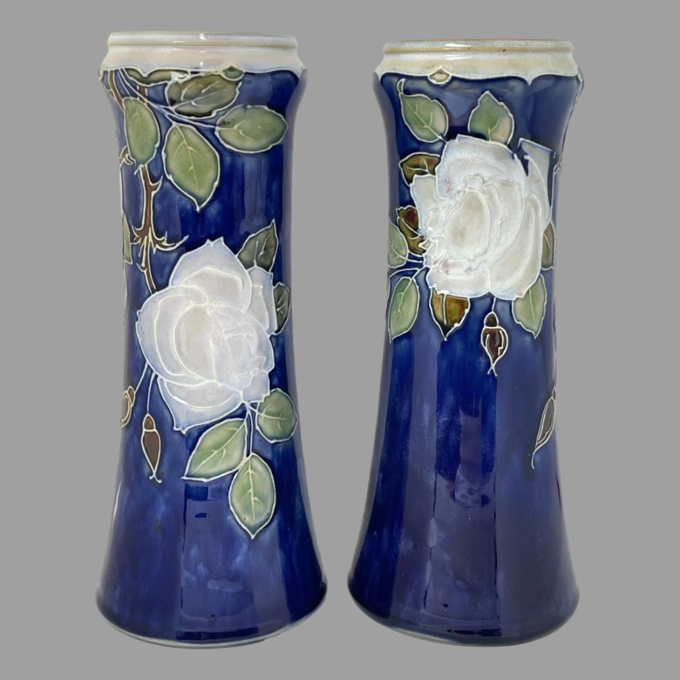 Main photo of Tall Doulton rose vases seen side by side from an eye level. The vase on left is displaying the side with rose lower down and the one on right has rose at the top. The background colour of the vases is a rich royal blue with one large off white rose to each with a few rose buds in a brown colour and leaves in a mixture of green and light brown. The rim of the vases is a creamy white. The vases can be displayed like this (one rose higher, one lower) or with both showing roses at same height. The vases have a lovely chimney-like shape and are stunning even without flowers!