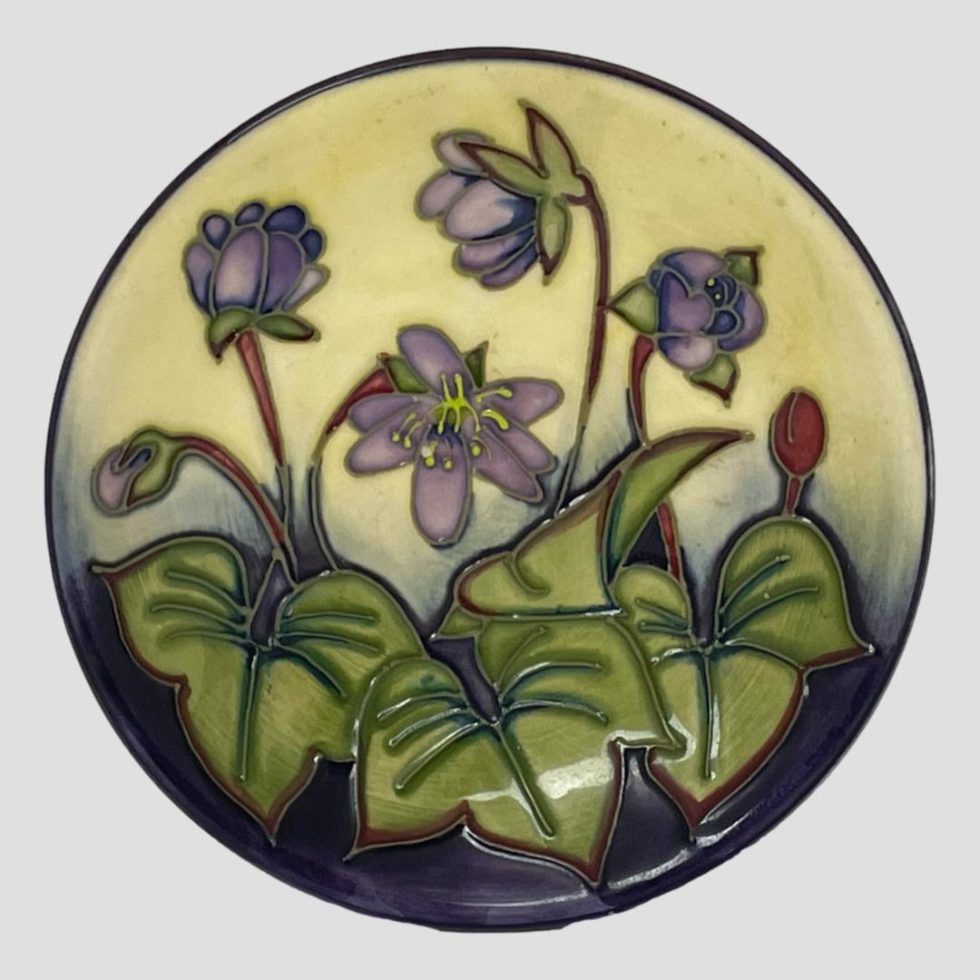 Main photo of Moorcroft Hepatica pin dish displayed upright with the hepatica flowers on full display. The flowers are a delicate lilac with wine red stalks whilst the leaves are a light green with dark green veins. The background colour is cream behind the flowers and dark purple behind the leaves.