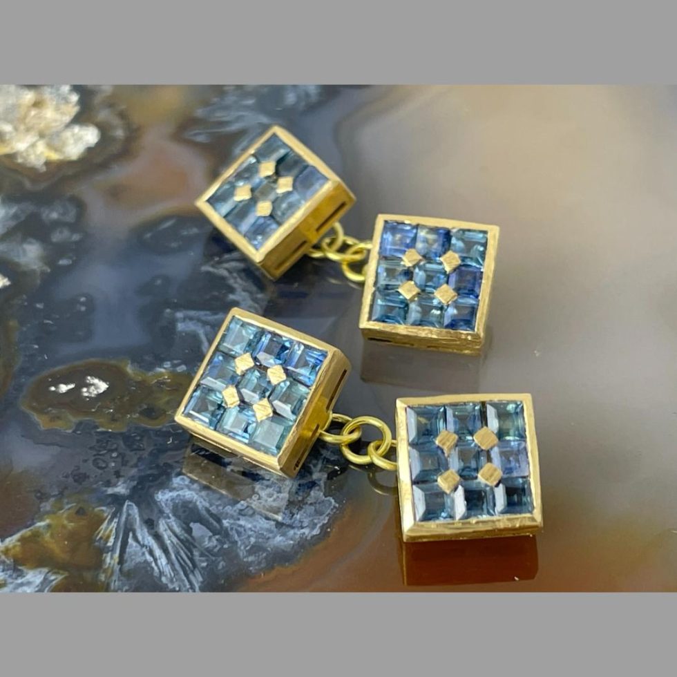 Photo of 18kt gold sapphire cufflinks displayed outside of their box and posed on top of some natural agate. Both sapphire ends of both cufflinks are on display with the chain divider visible in between the 2 ends. The square cut sapphires within a square frame of the cufflink ends is very appealing and there are 4 gold pin dividers to separate the sapphires.