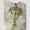 Main photo of Edwardian gold peridot pendant shown displayed on top of a rock of quartz crystal. The bail which looks a little like a closed fan has a seed pearl to its centre. The main body of the pendant has at its widest area at the top with a row of foliage finishing with a flower to the centre. Below this area there is an inner circular frame with seed pearls at each compass point and a round cut peridot to the centre. Another seed pearl is found below this area followed by the larger of the 2 round cut peridot stones which is free hanging.