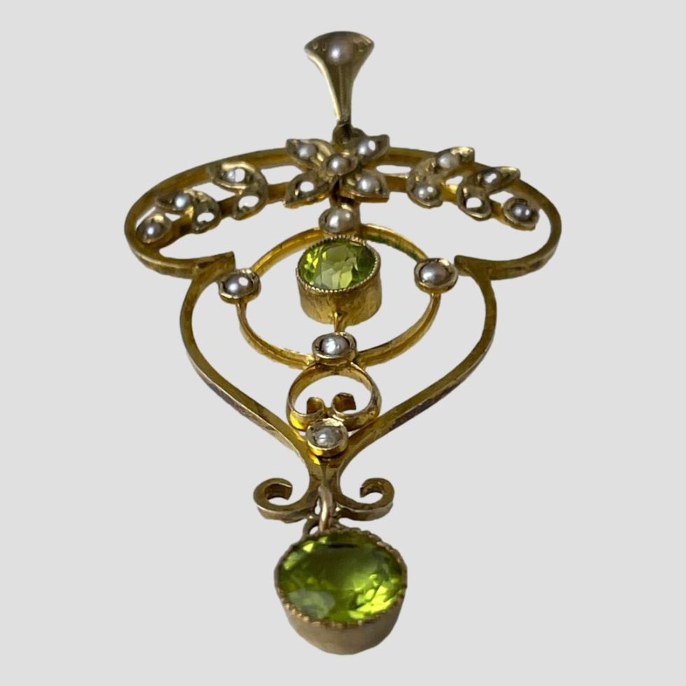 Photo of Edwardian gold peridot pendant shown laid on a flat surface. The larger peridot stone at the bottom of the pendant is in the centre foreground tapering away to the bail in the centre back.